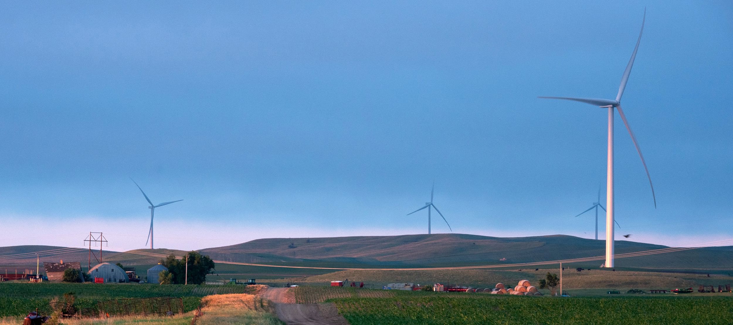 Rolling farmland with barns and farm equipment surrounded by four wind turbines
