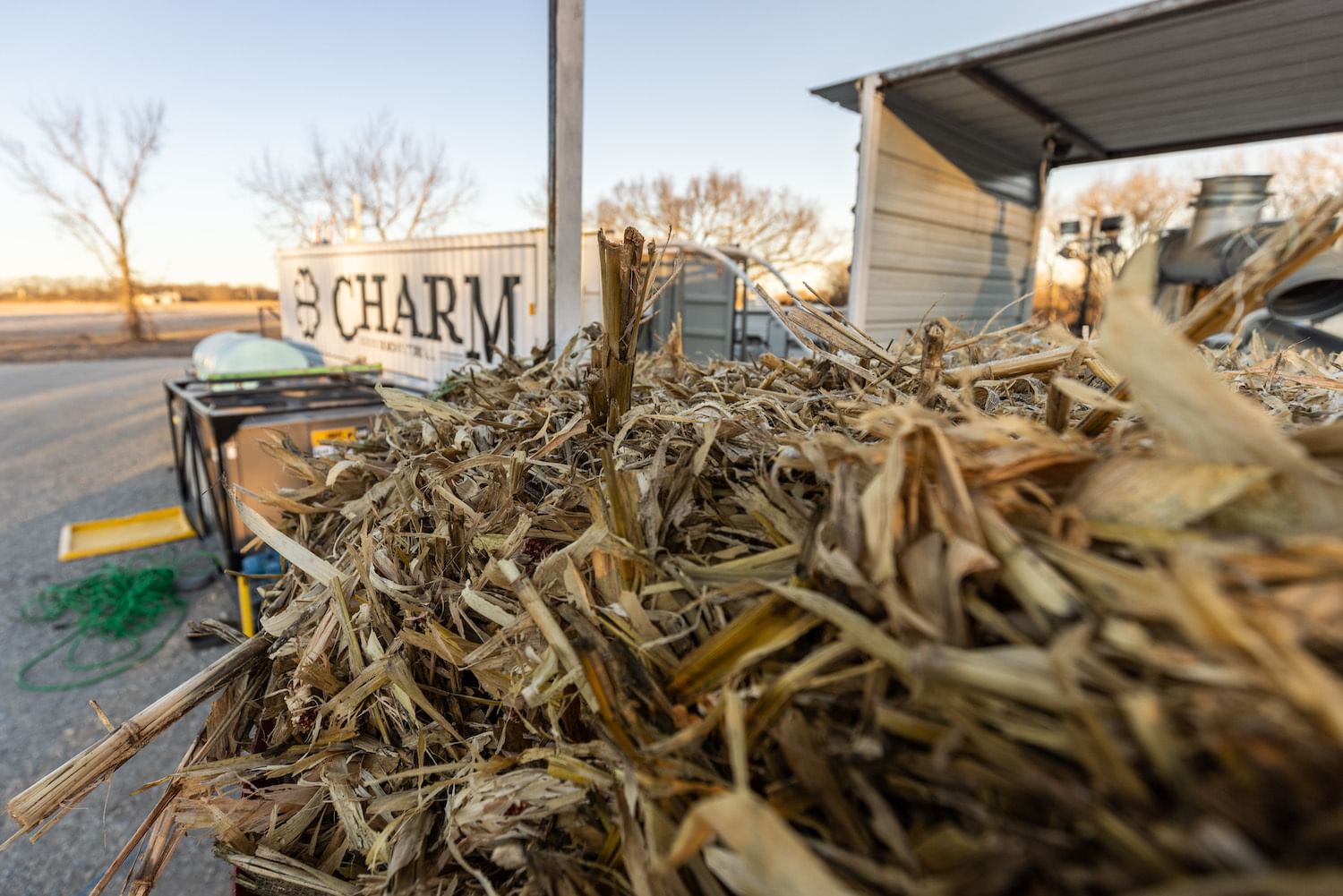 A pile of dried corn leaves is shown in the foreground, near a white shipping container with the word "Charm" on the side
