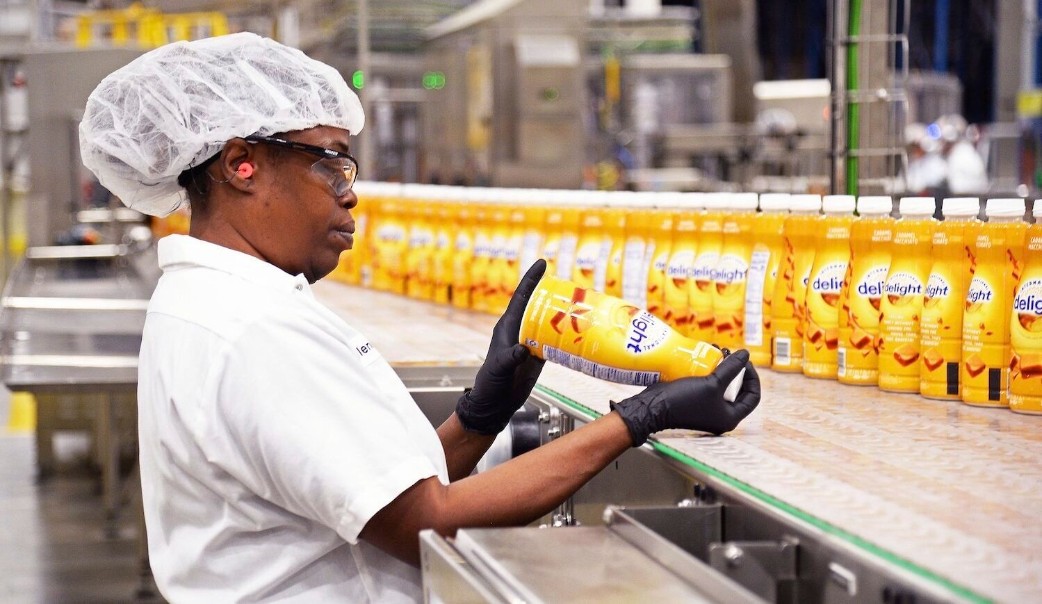 A woman in white shirt and white hair net examines yellow bottles of Delight on a factory line