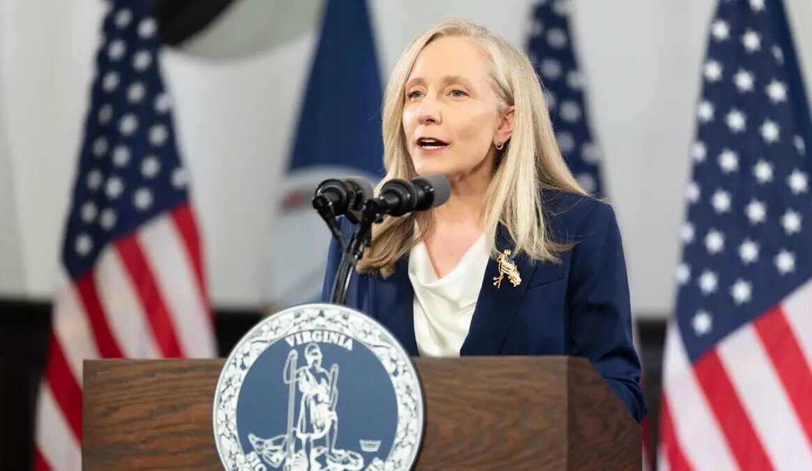 A woman in business attire speaks into a mic at a podium in front of U.S. flags