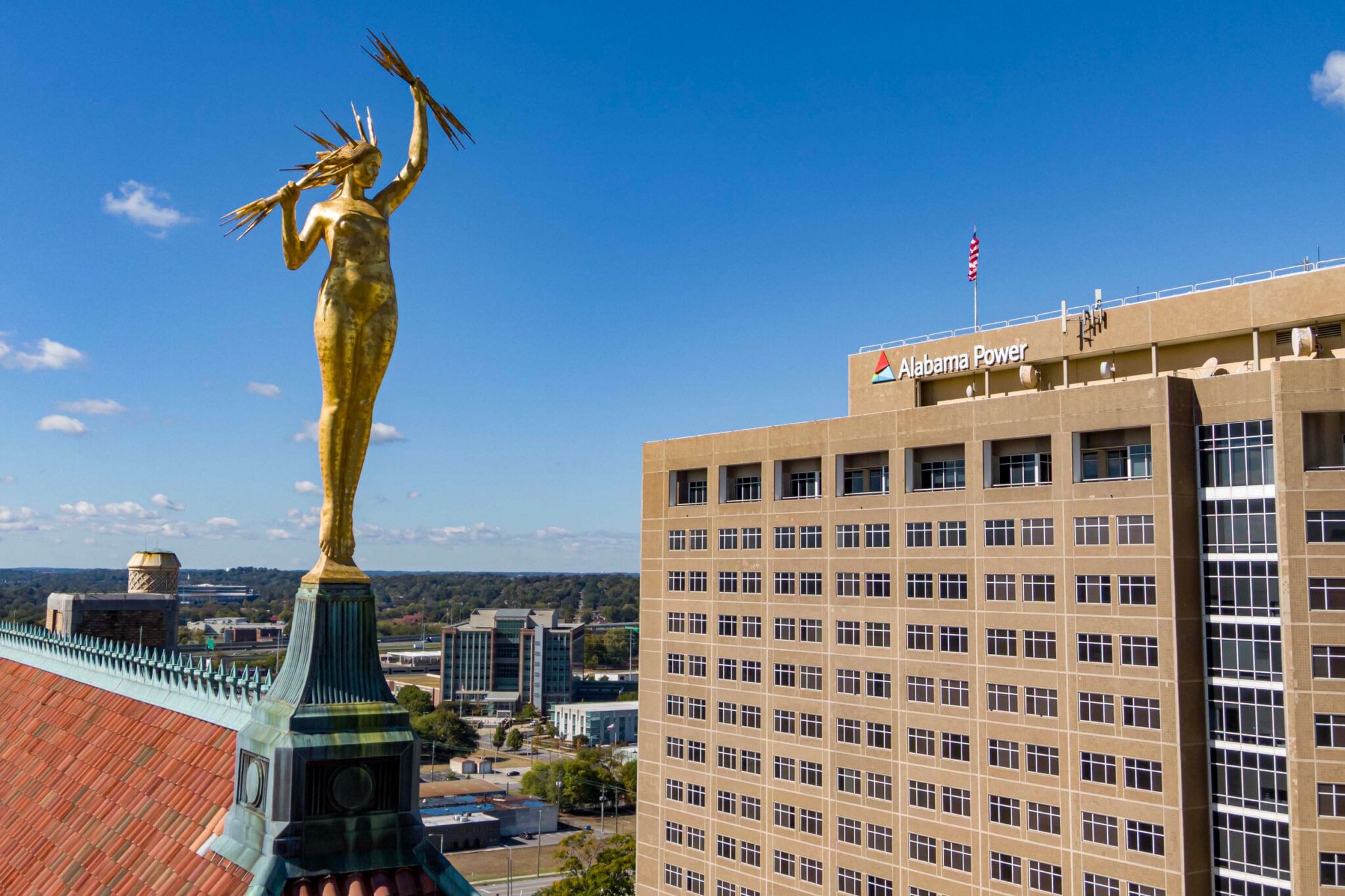 Golden statue poised on a roof, shot from above, with a brown high-rise with an "Alabama Power" sign and a US flag next door