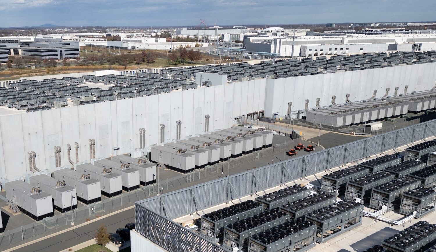 Large warehouses flanked by a dozen generators on a lot, with the rooftop fans on another building in the foreground