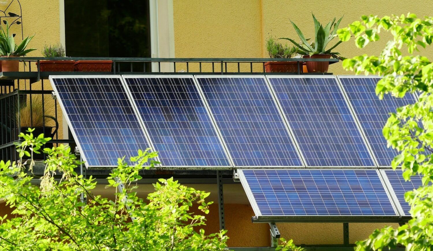 Solar panels on a balcony of a home with yellow walls, plants on balcony, and trees in front