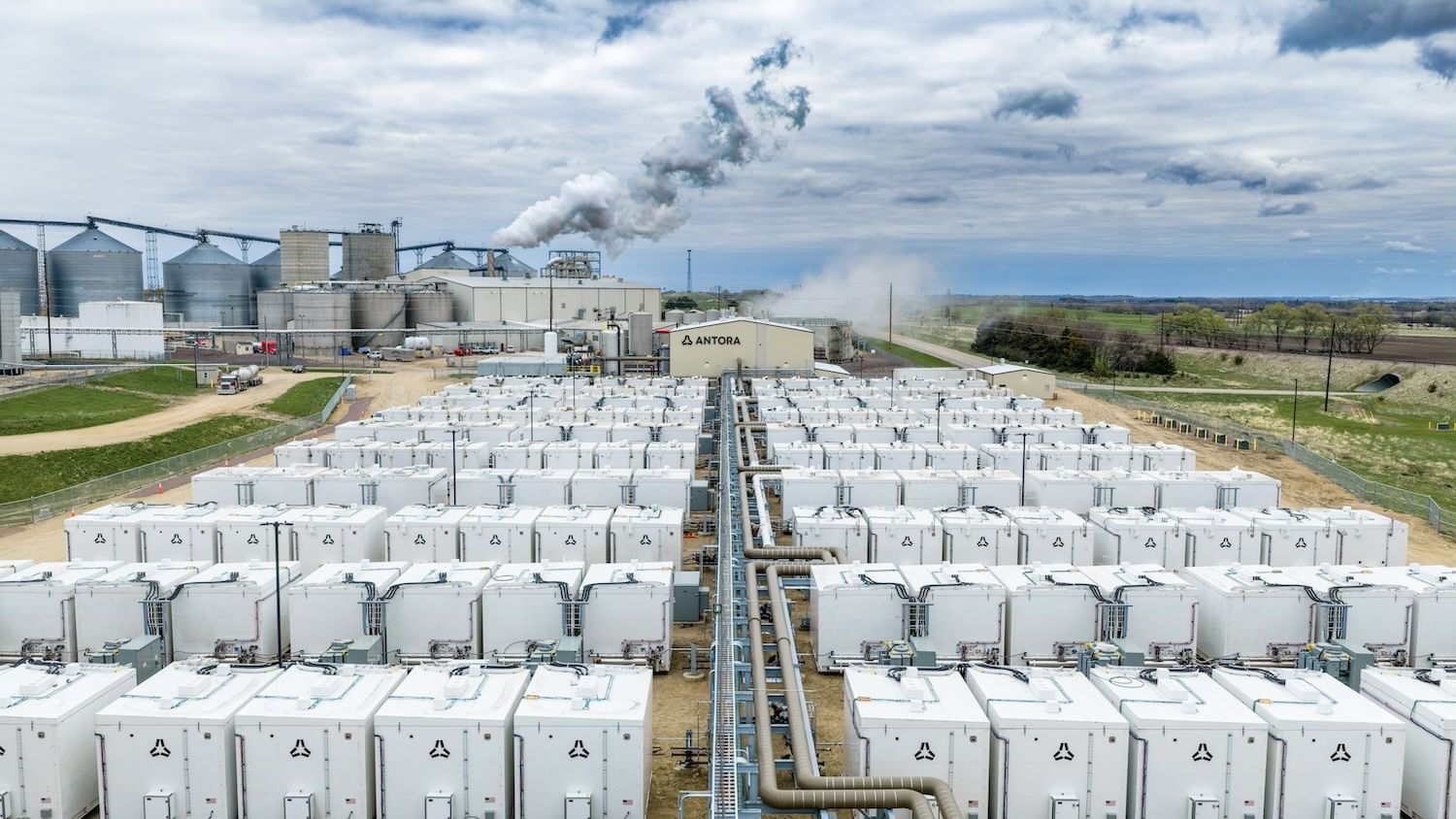 Rows of white batteries in front of a factory, with a beige building between with an Antora sign
