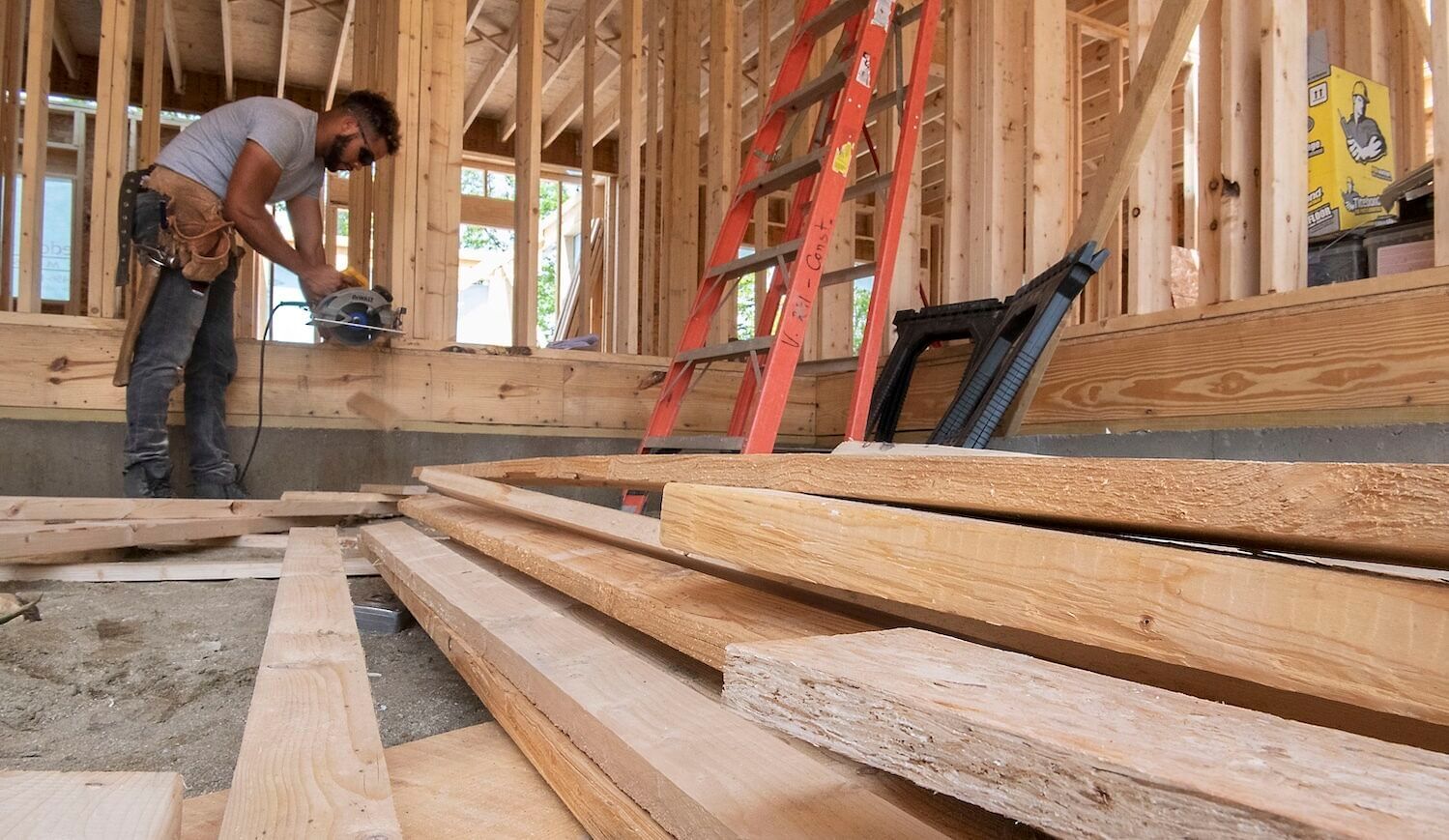 A person holding a circular saw bends over a beam of wood in a half-finished home
