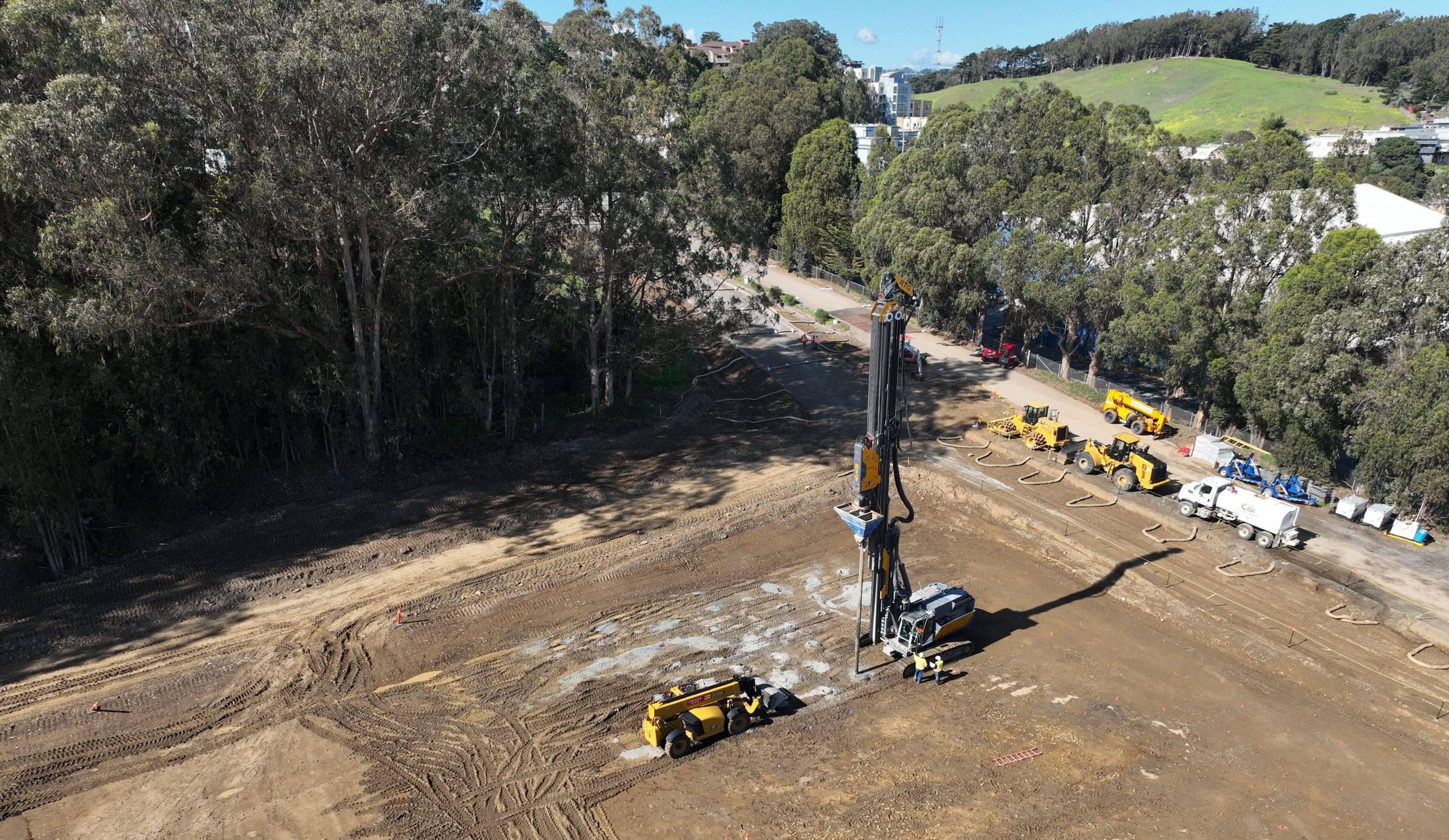 Dirt lot being leveled, with a view of a hilly park, McClaren, and Coit Tower far in the distance