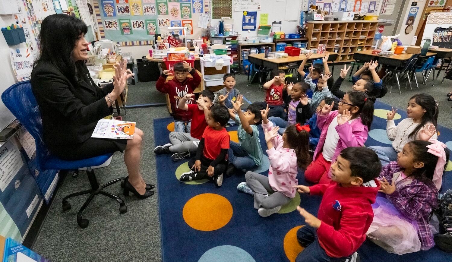 A group of young kids sitting on a colorful rug in a classroom while a woman sits on a chair holding a book.