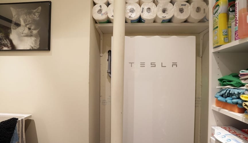 A photo of a cat hanging on a white wall next to a Tesla battery, with packages of paper towels on a shelf above it