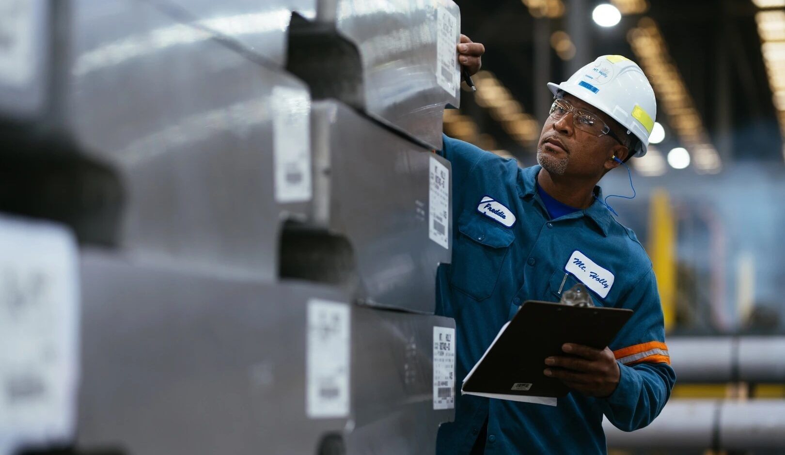 A man in a white hard hat with a blue worksuit inspects rolls of aluminum