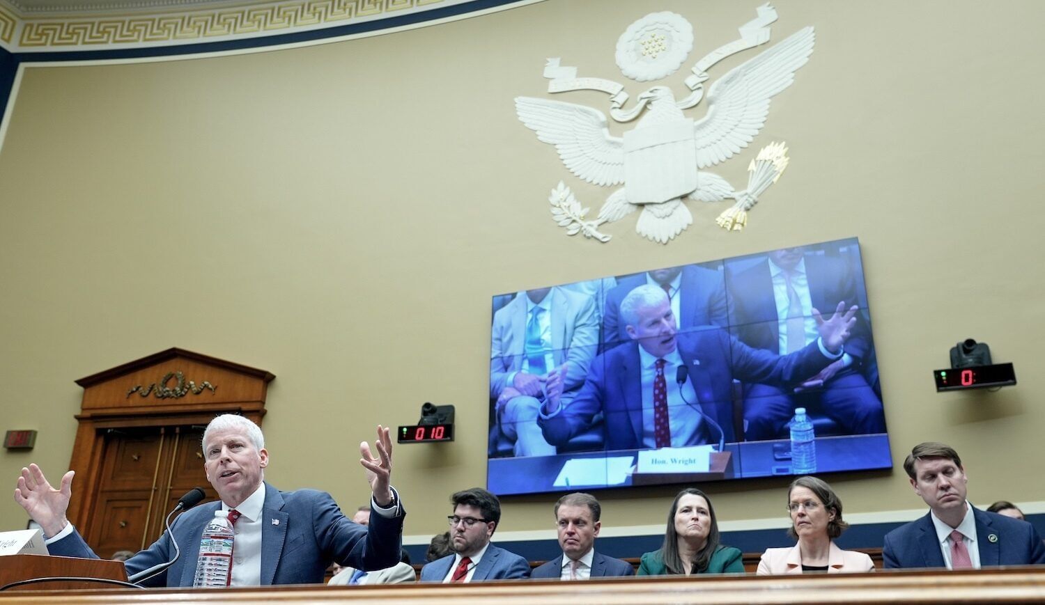 Chris Wright talking with his arms in the air; an image of him is on a large screen on the wall behind him