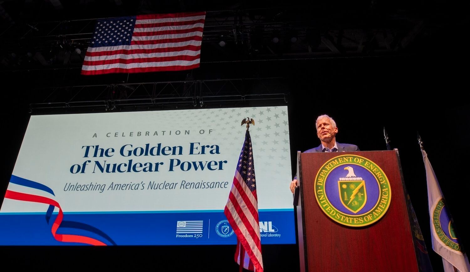 Man at a podium with sign saying "A Celebration of the Golden Era of Nuclear Power" and several flags