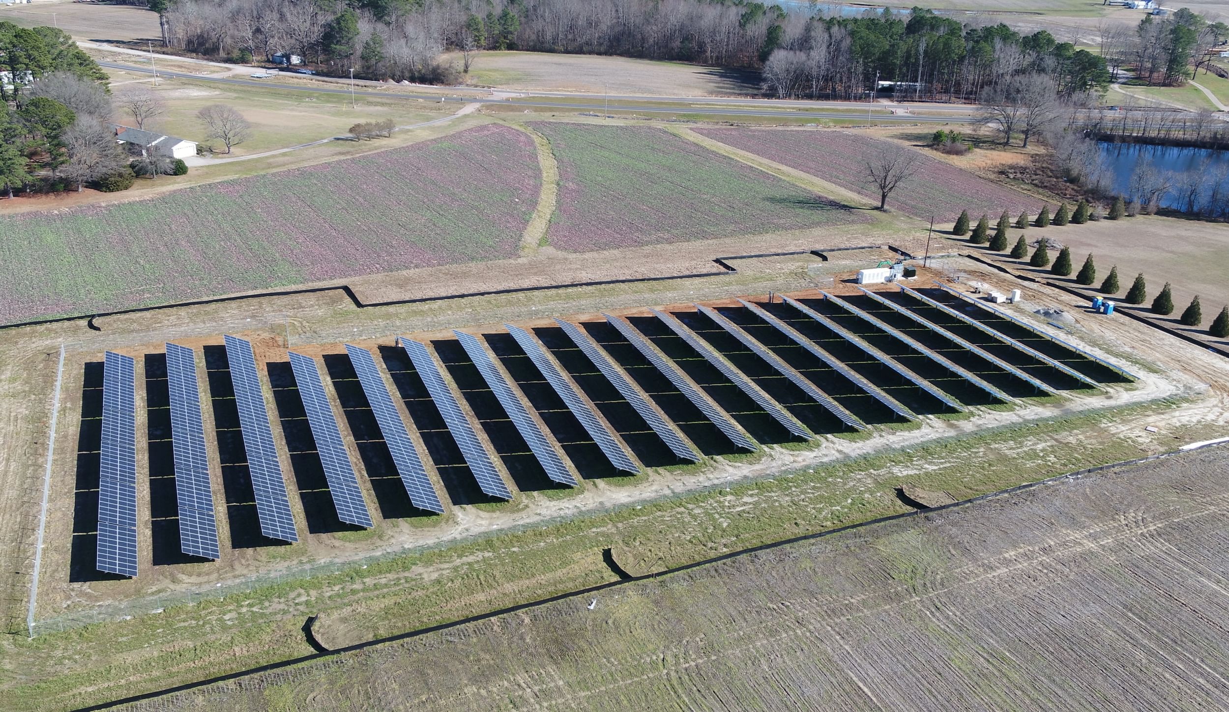 Solar panels on a field with a battery surrounded by trees, a lake, and what looks like farmland
