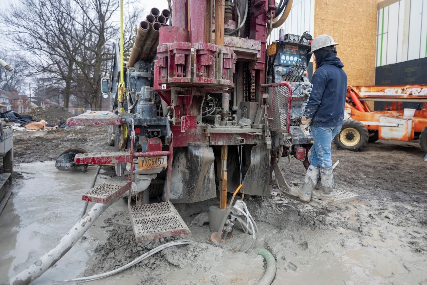 Worker in jeans and blue jacket, hard hat, operates a red drilling rig on muddy ground