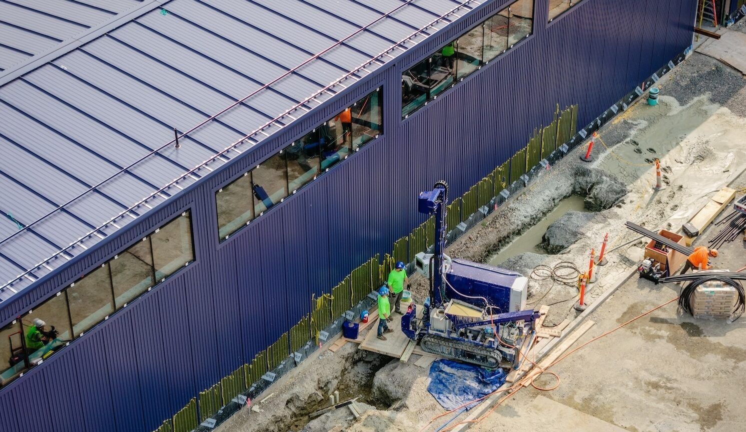 Overhead shot of a blue-sided and -roofed building with a compact drilling rig along side it and a few workers