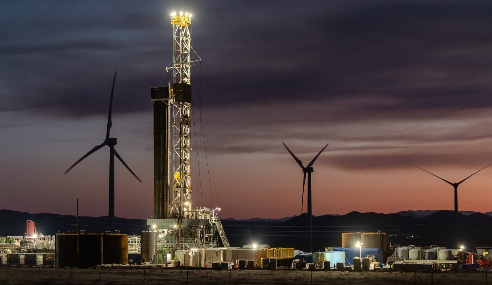 Geothermal plant with lights on at dusk or dawn with pink/orange sky and three wind turbines in background