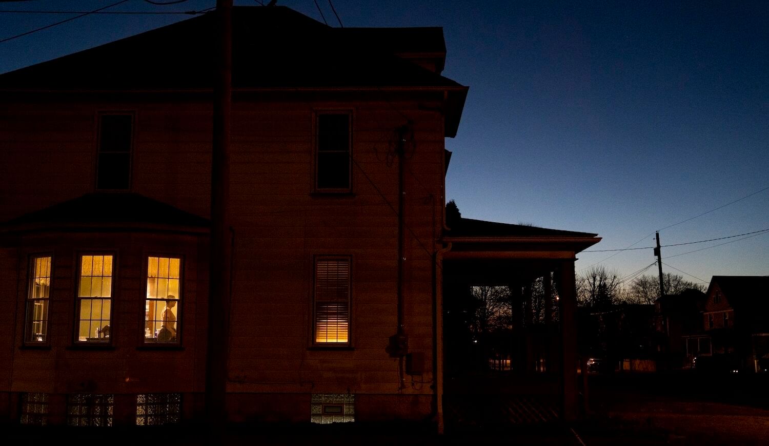 Two-story house with three windows fully lit on the bottom floor and a person seen walking inside a room through one of them