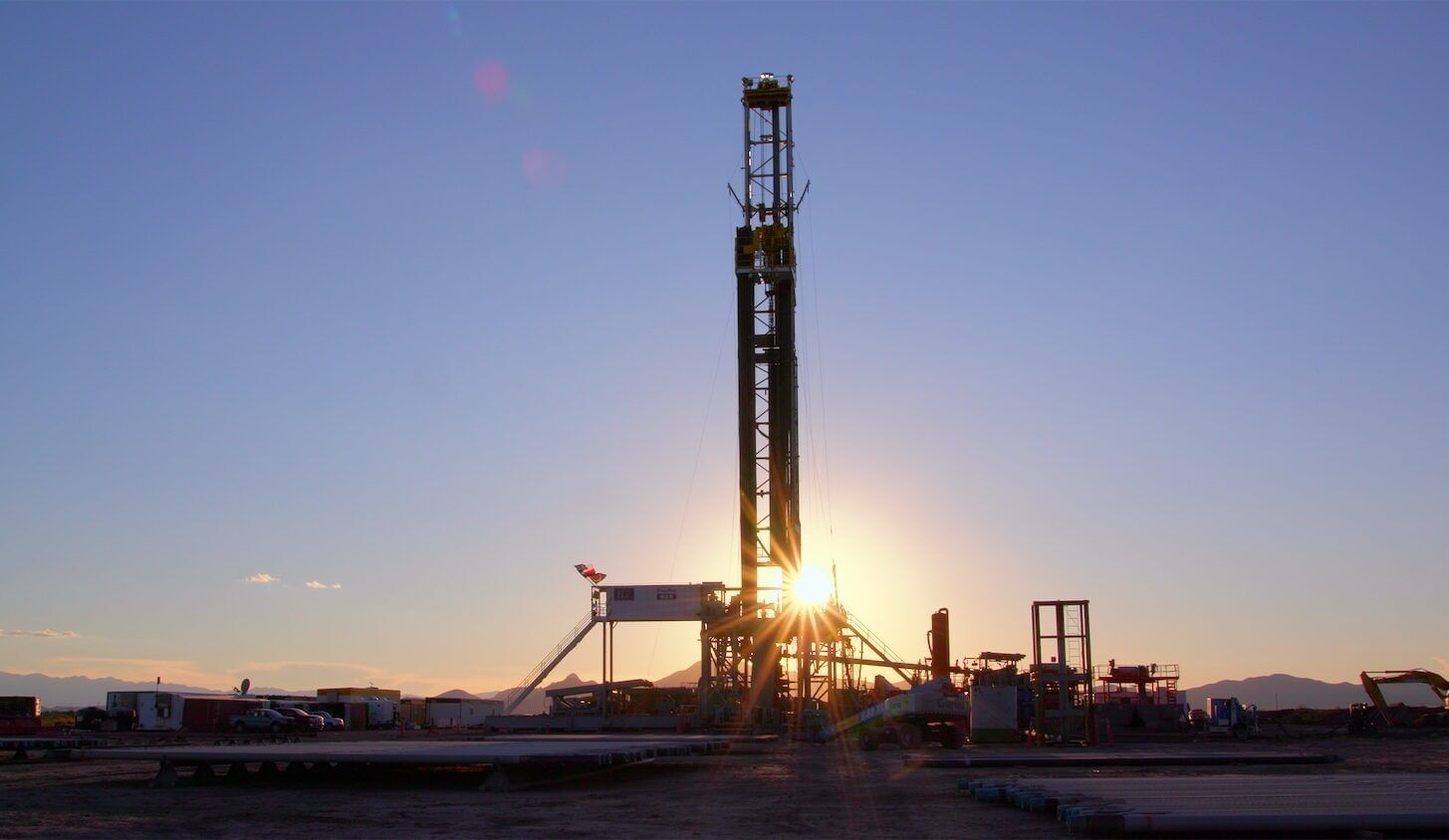 A large drill crane with the sun shining through it near dusk or dawn, surrounded by various equipment