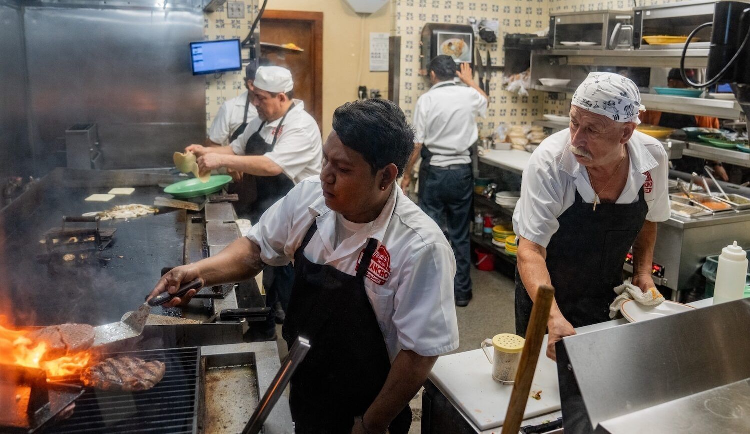 Five white-shirted and dark-aproned workers in a kitchen with an open flame and long cooktop
