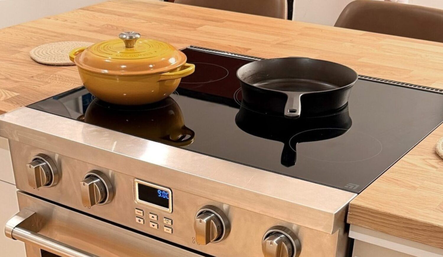 An induction stovetop with a yellow Le Creuset and a cast-iron pan on it; surrounded by a wood countertop with 2 pot holders
