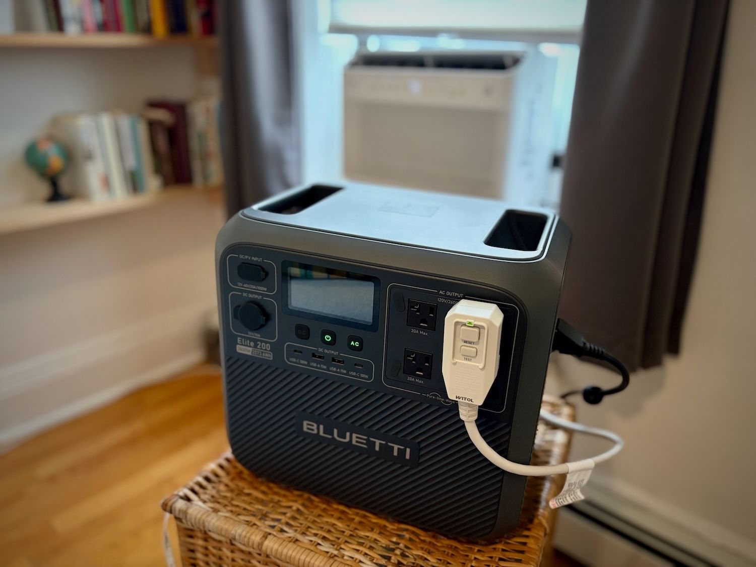 Black battery plugged into a window AC unit atop a wicker hamper