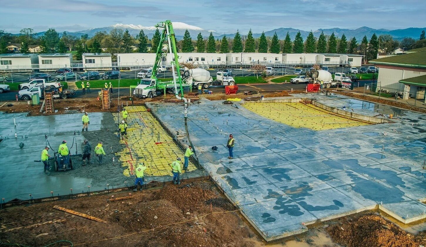 Concrete being poured by numerous workers with buildings, trees, and mountain in background