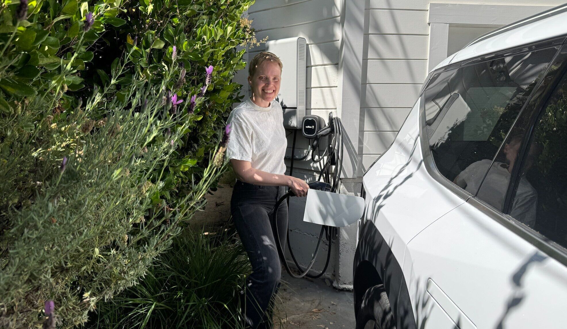 A woman in a white shirt and blak pants smiles next to a white car and black charging cord on white home; plants on the left