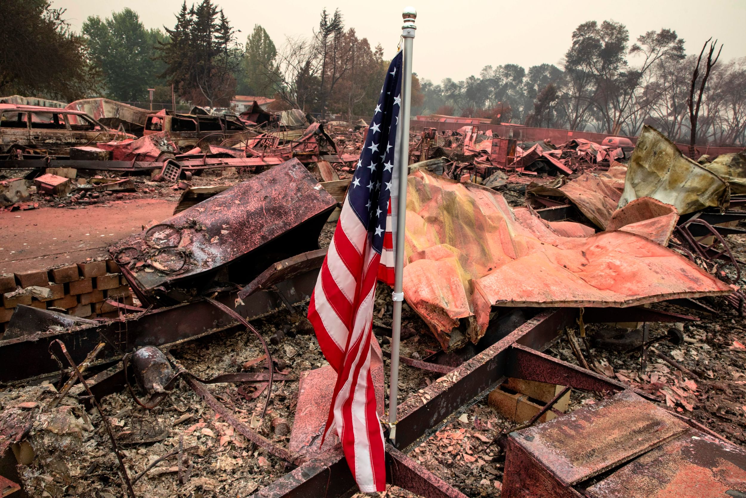 An American flag in hunks of twisted, burnt metal behind it and a backdrop of trees and smoky sky