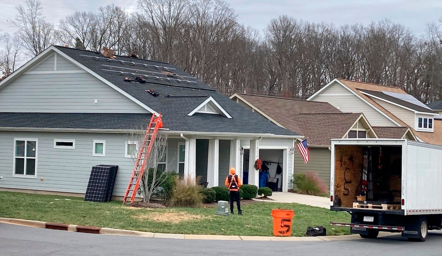 Solar panels lean on the side of a house. A worker stands in the house's yard.