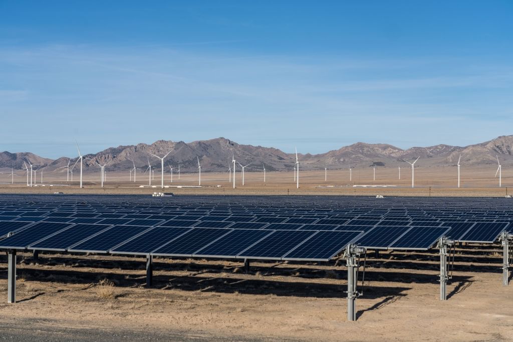 A solar panel array and a collection of wind turbines sit on brown, desert-like ground.