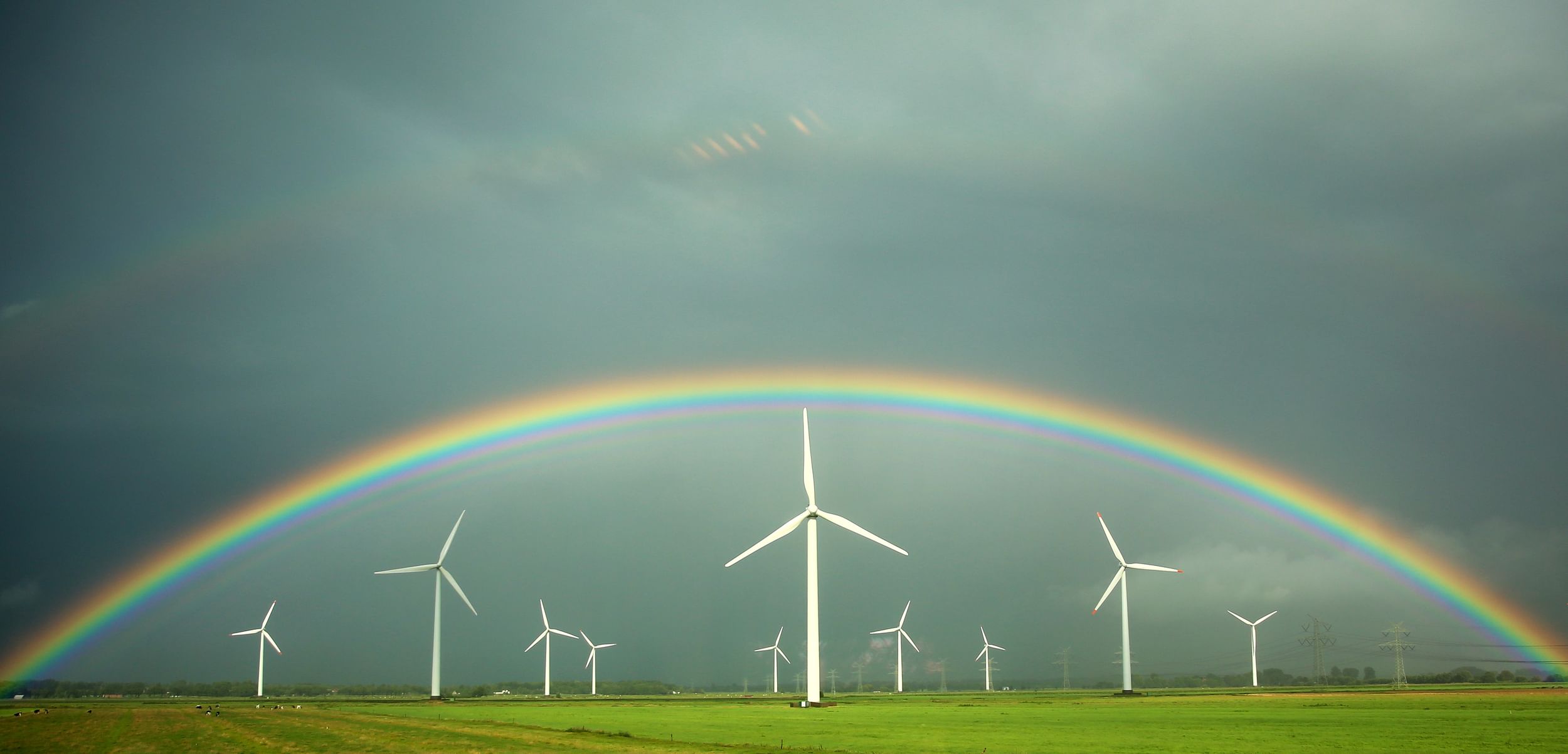 Green field with 10 wind turbines in the distance, a rainbow arching above them in a stormy grey sky
