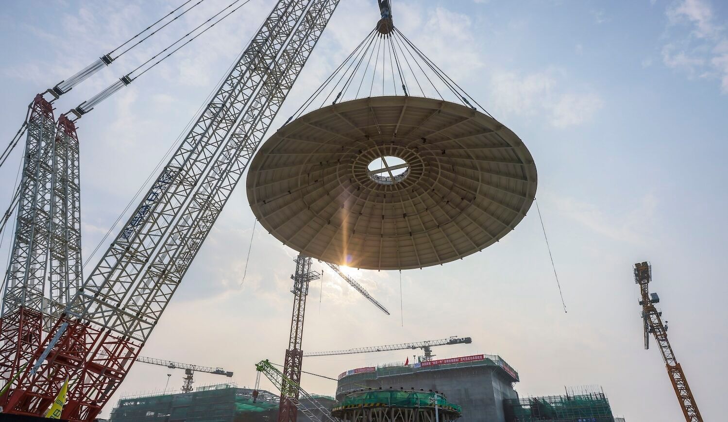 A large crane holds a dome structure against a blue sky, with industrial equipment in the background.