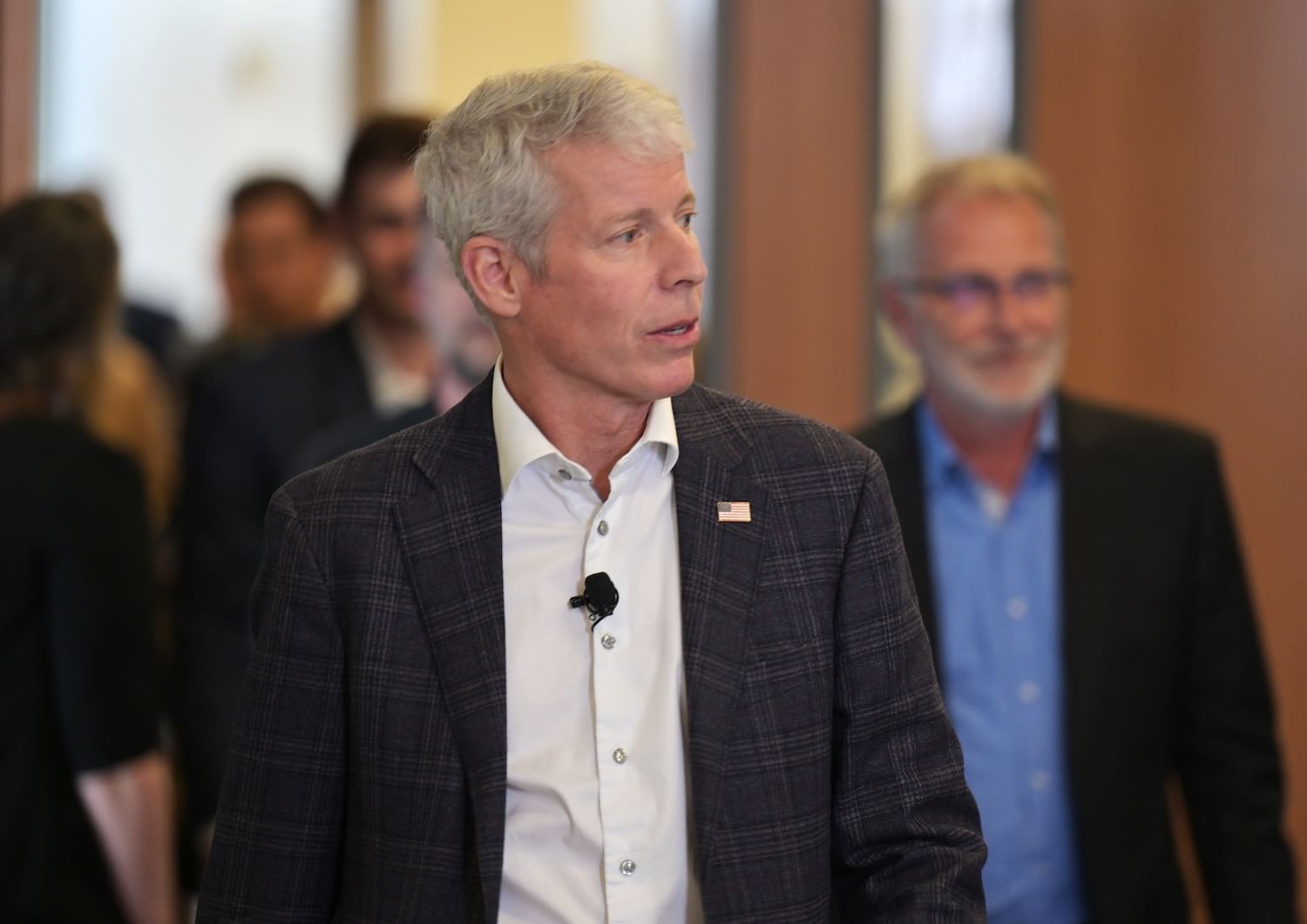 A person wearing a suit and an American flag pin walks in a room. Blurred people are behind him.