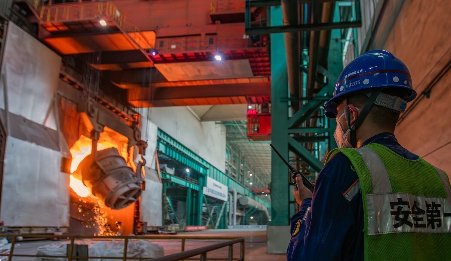 A person wearing a hard hat and work vest holds a walkie talkie and stands in front of industrial operations