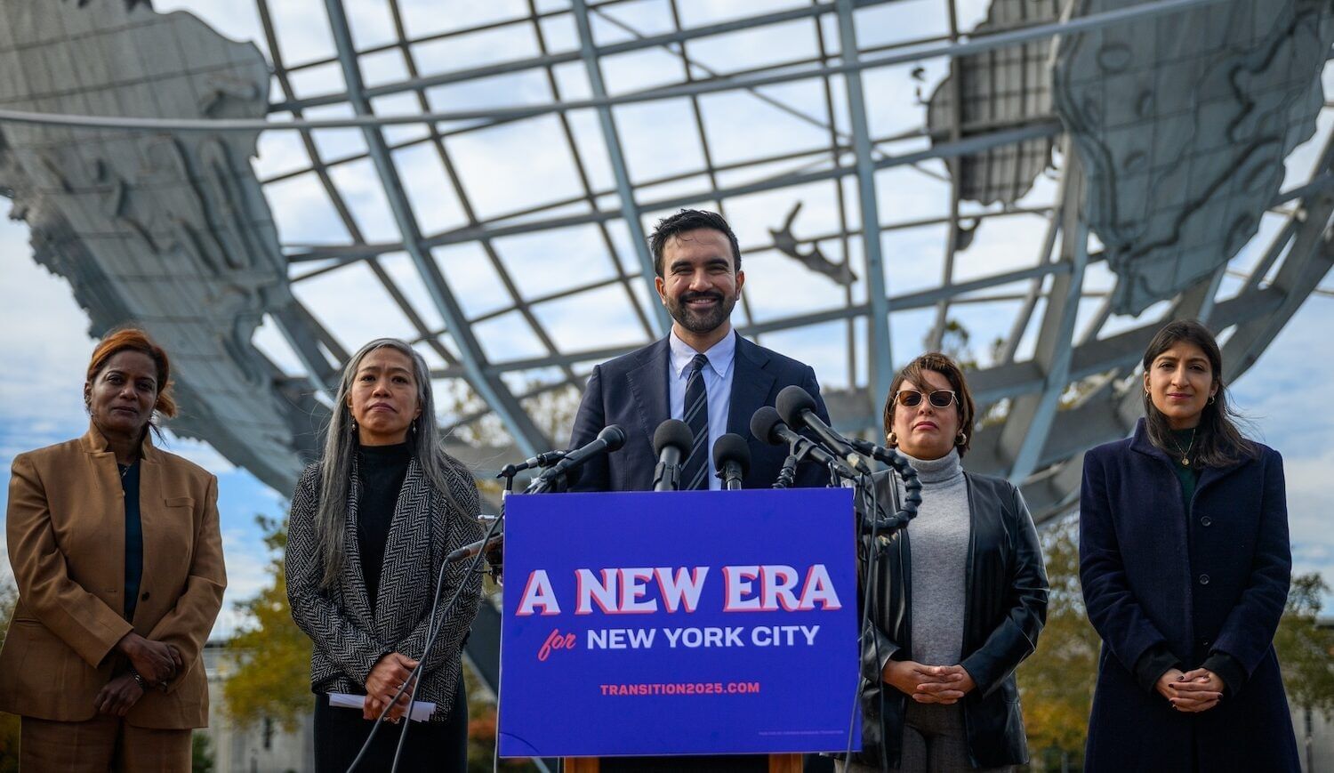 A man at a podium that reads "A New Era New York City." He is flanked by four people, standing in front of a large globe.