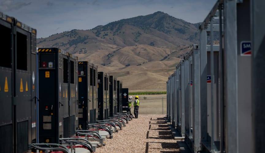 Two rows of battery storage units, with two workers at the end of a row, mountains in distance