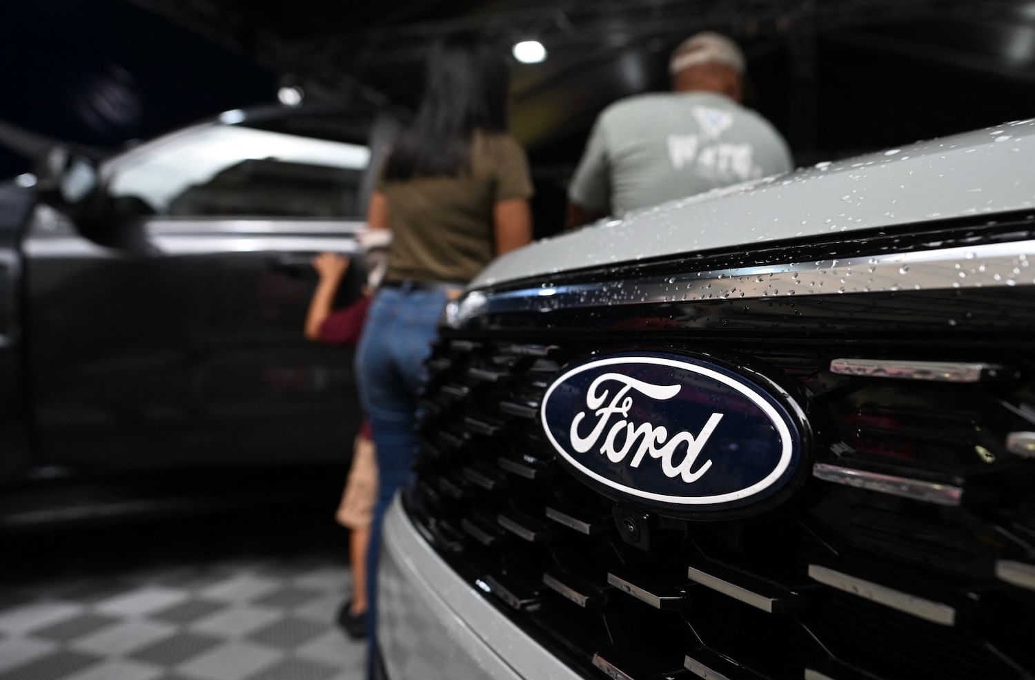 A logo of the U.S. Ford Motor Company on the front grille of a truck at an auto show with show, with a few people behind it
