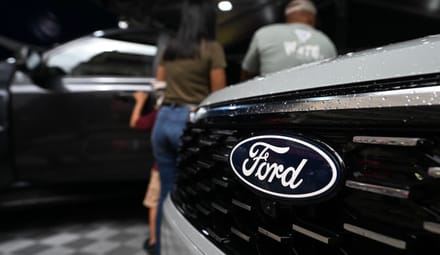 A logo of the U.S. Ford Motor Company on the front grille of a truck at an auto show with show, with a few people behind it