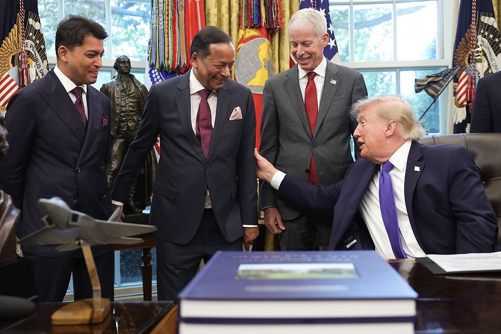 Three men in suits standing, all smiling, next to Trump sitting at a desk, with lots of flags behind them