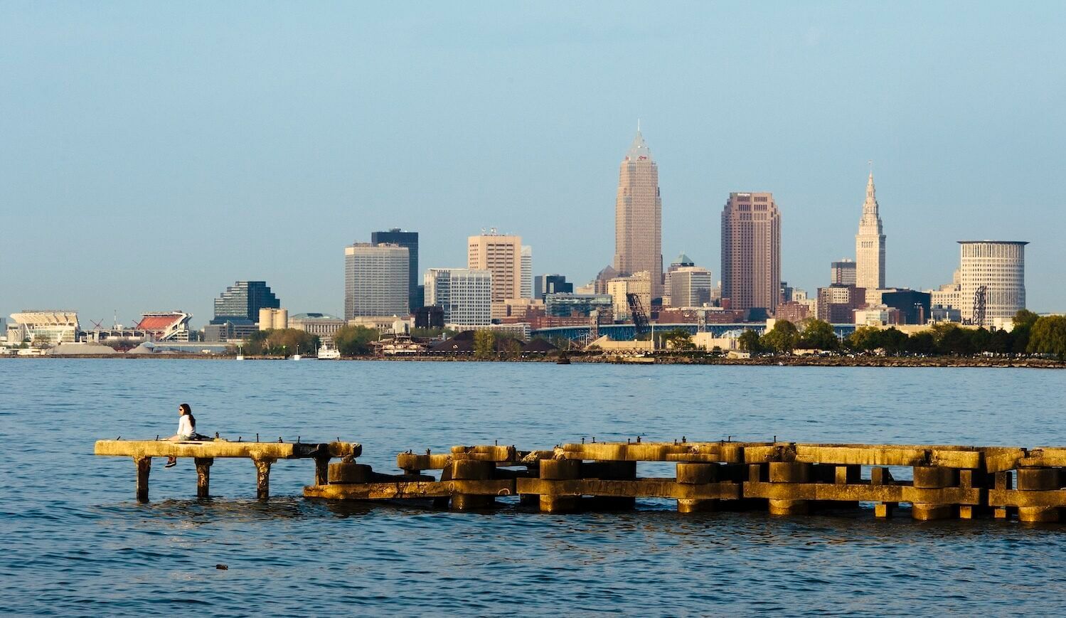 A woman sits on a pier in the water in front of a city skyline.