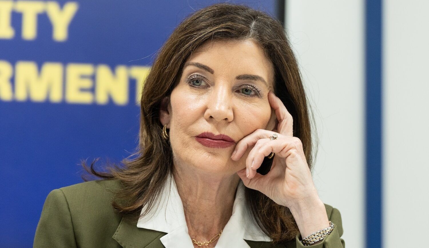Woman with brown hair in an olive-green suit and white shirt, left hand on face