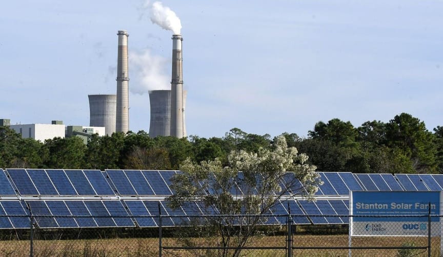A row of solar panels in front of a row of trees with smokestacks behind
