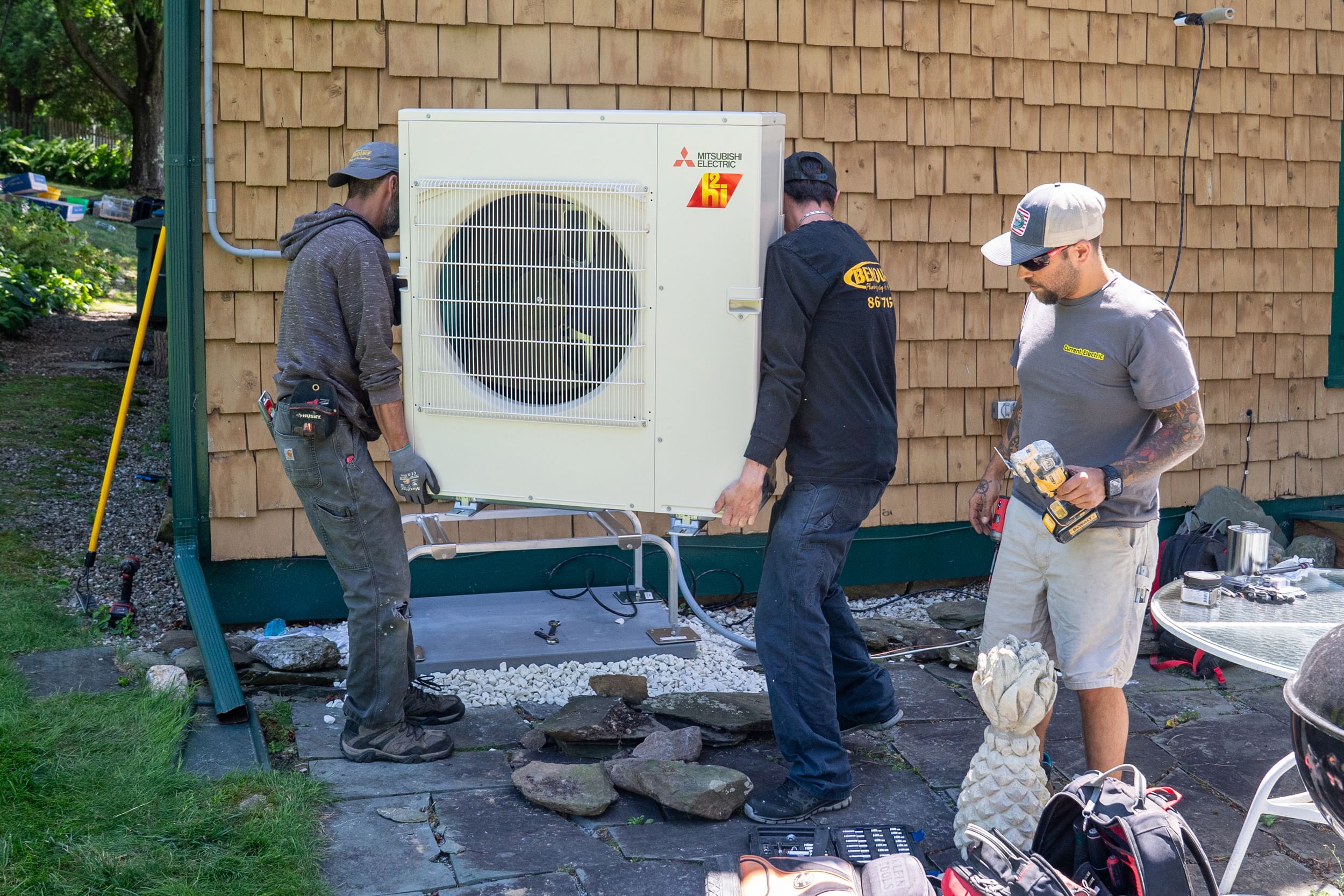 Two men carrying a heat pump by a shingled house, while a third holds a tool