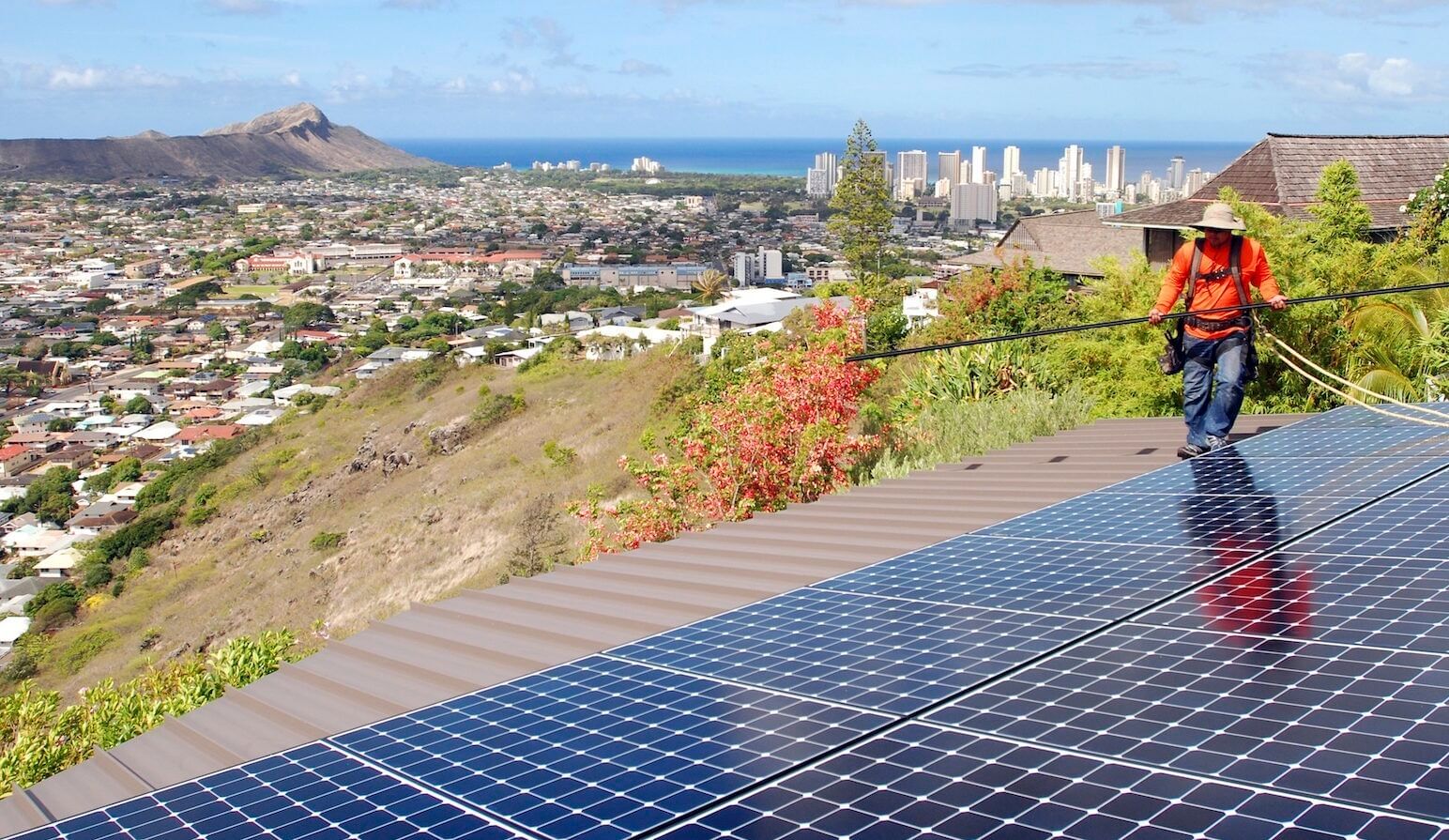 Worker in an orange shirt near a solar array with beautiful views of the city, Diamond Head, and the ocean