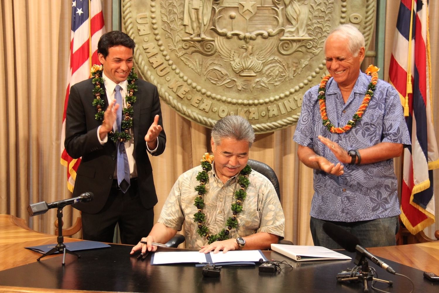 Three men, two standing and clapping; man in center with pen and document; state seal and two flags behind them