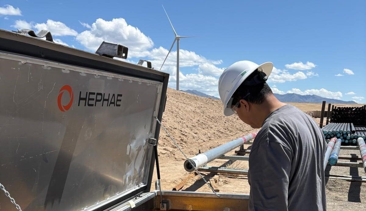 Man in white hard hat and gray shirt by gray container, with long pipe and wind turbine visible