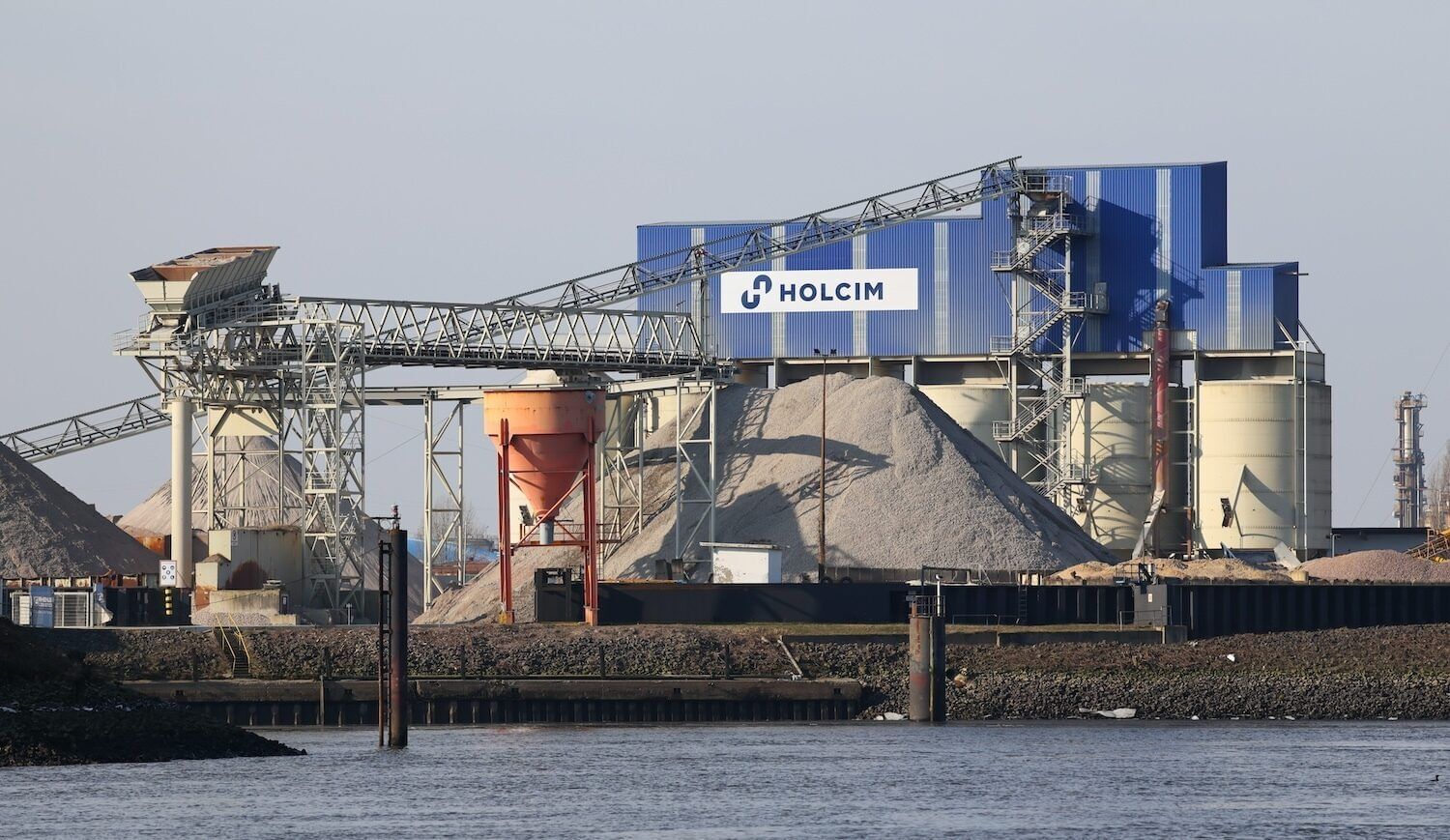 Blue-sided building with Holcim name and logo above cement-making supplies and equipment; a body of water in the foreground