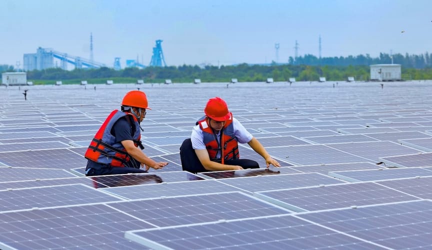 Two men in red hats and vests amid solar panels, with a coal facility in the distance