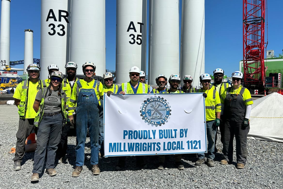 A group of Millwrights Local 1121 workers in hard hats stand in front of wine turbine towers.