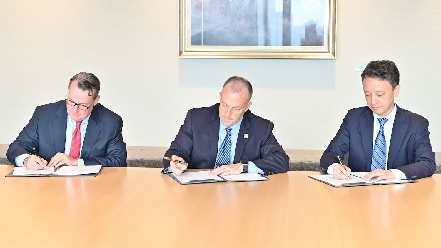 Three men in suits seated at a light-wood table, heads down signing documents