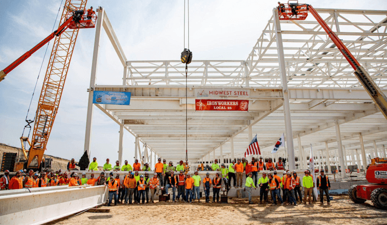 A large group of people in safety vests and hardhats stand next to an under-construction factory